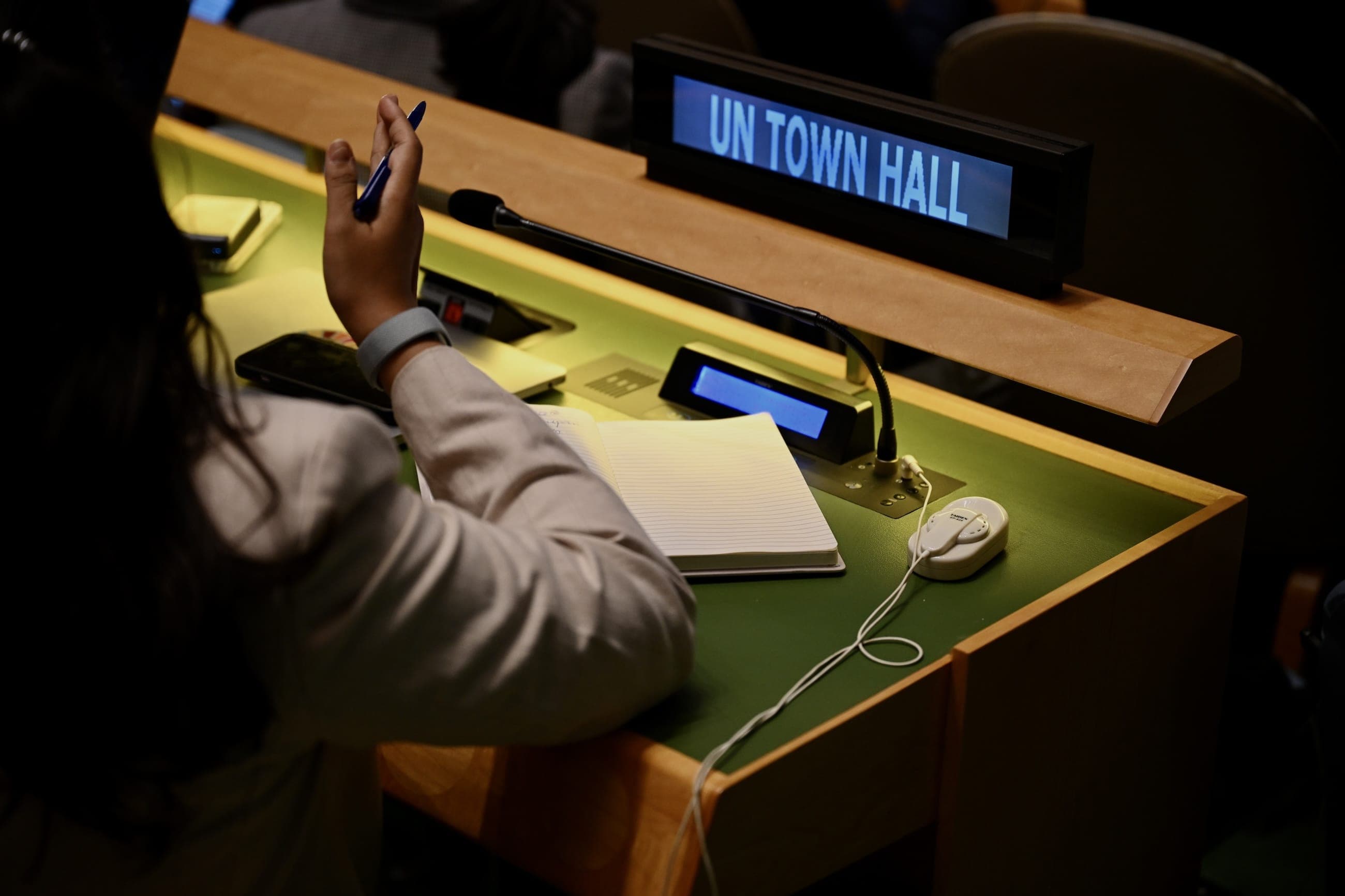 An attendee raises a pen beside a United Nations town hall desk nameplate.