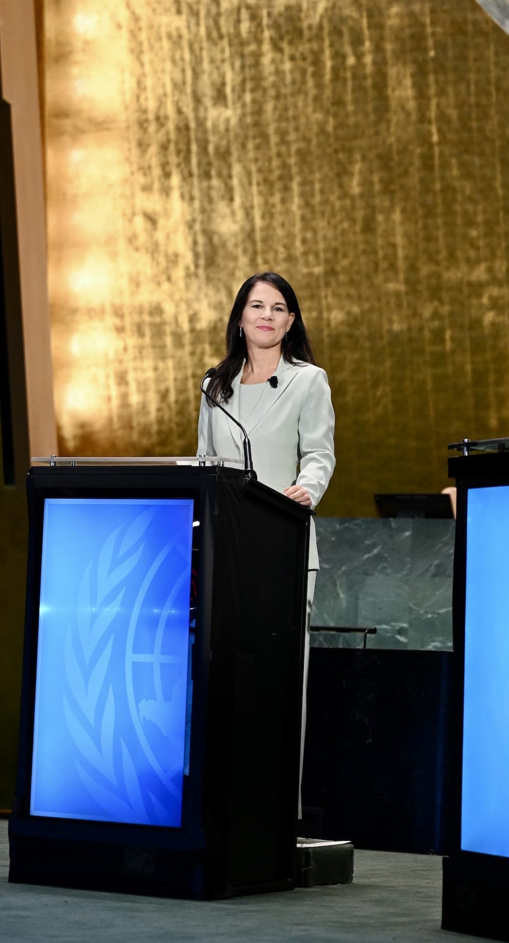 A woman in a light suit stands at a United Nations lectern before the General Assembly Hall’s gold wall.