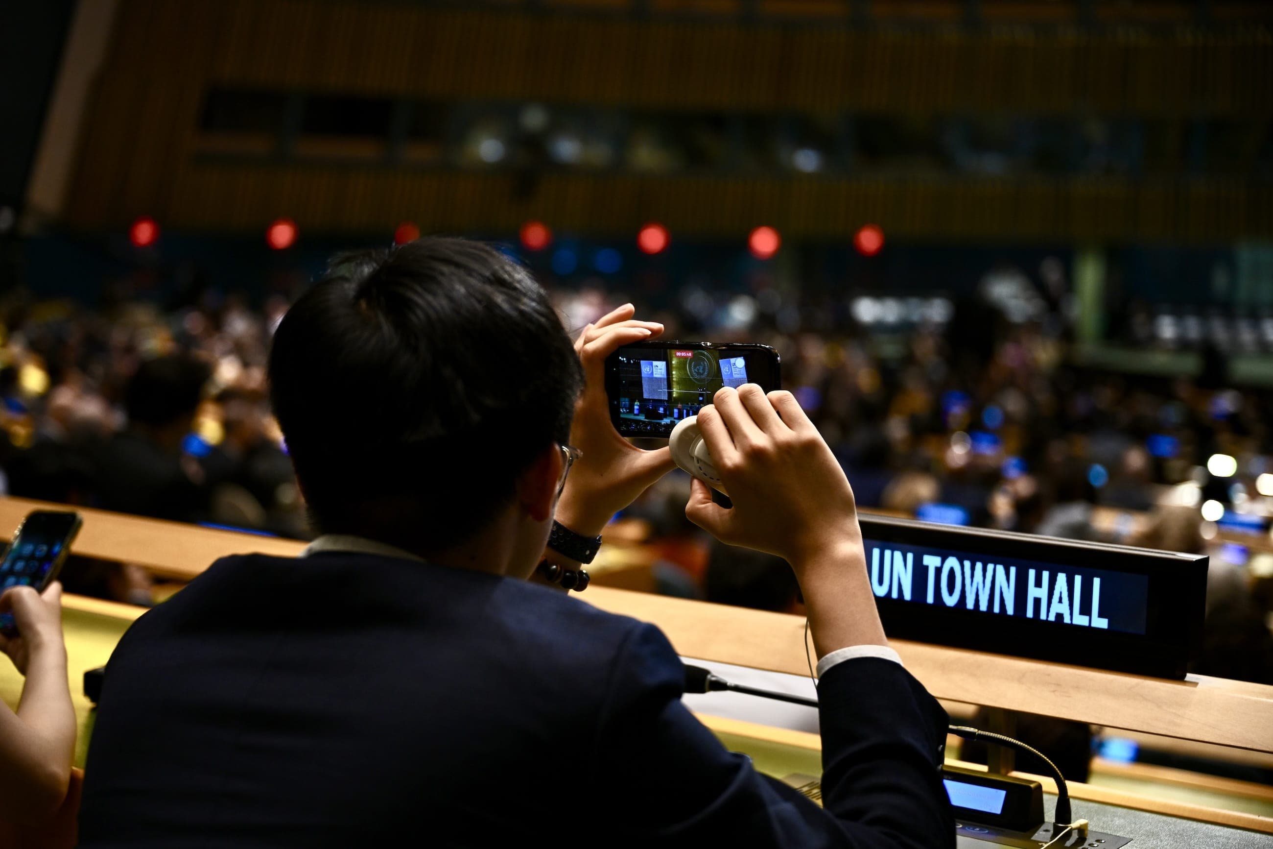 Delegates watch a speaker on a large screen in a United Nations conference room.