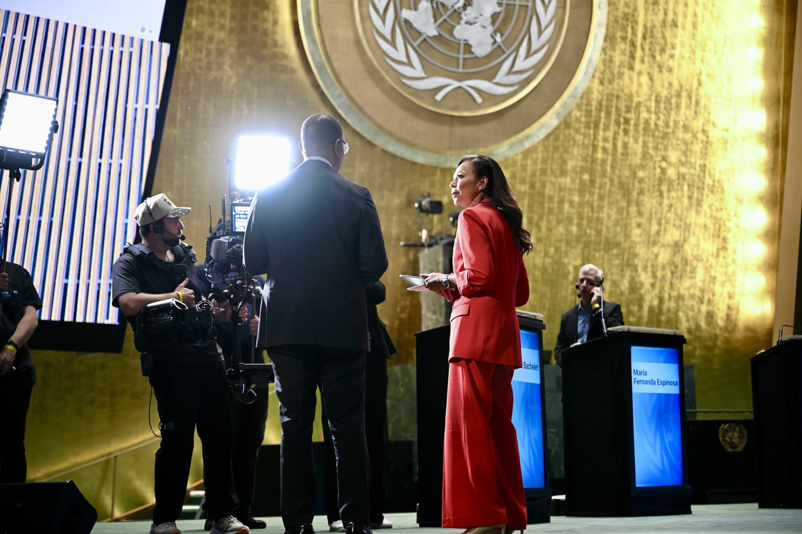 A woman in a red suit stands on the General Assembly Hall stage beside a camera crew.
