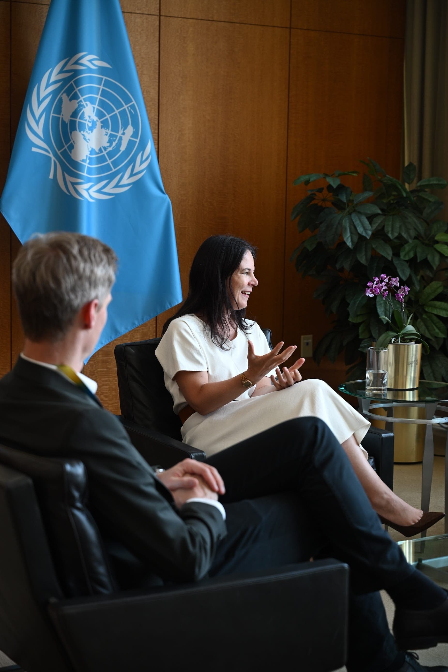 A woman in a white dress speaks during a seated discussion before a United Nations flag.