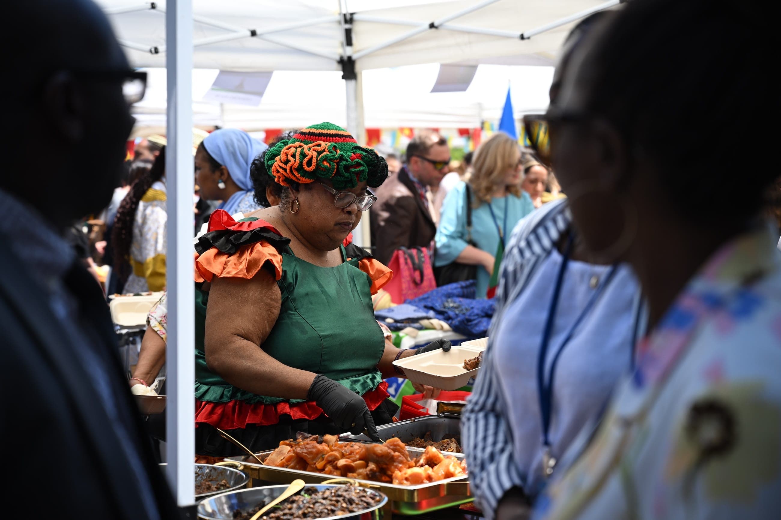 A woman in a green dress and colourful headwrap serves food at an outdoor cultural event.