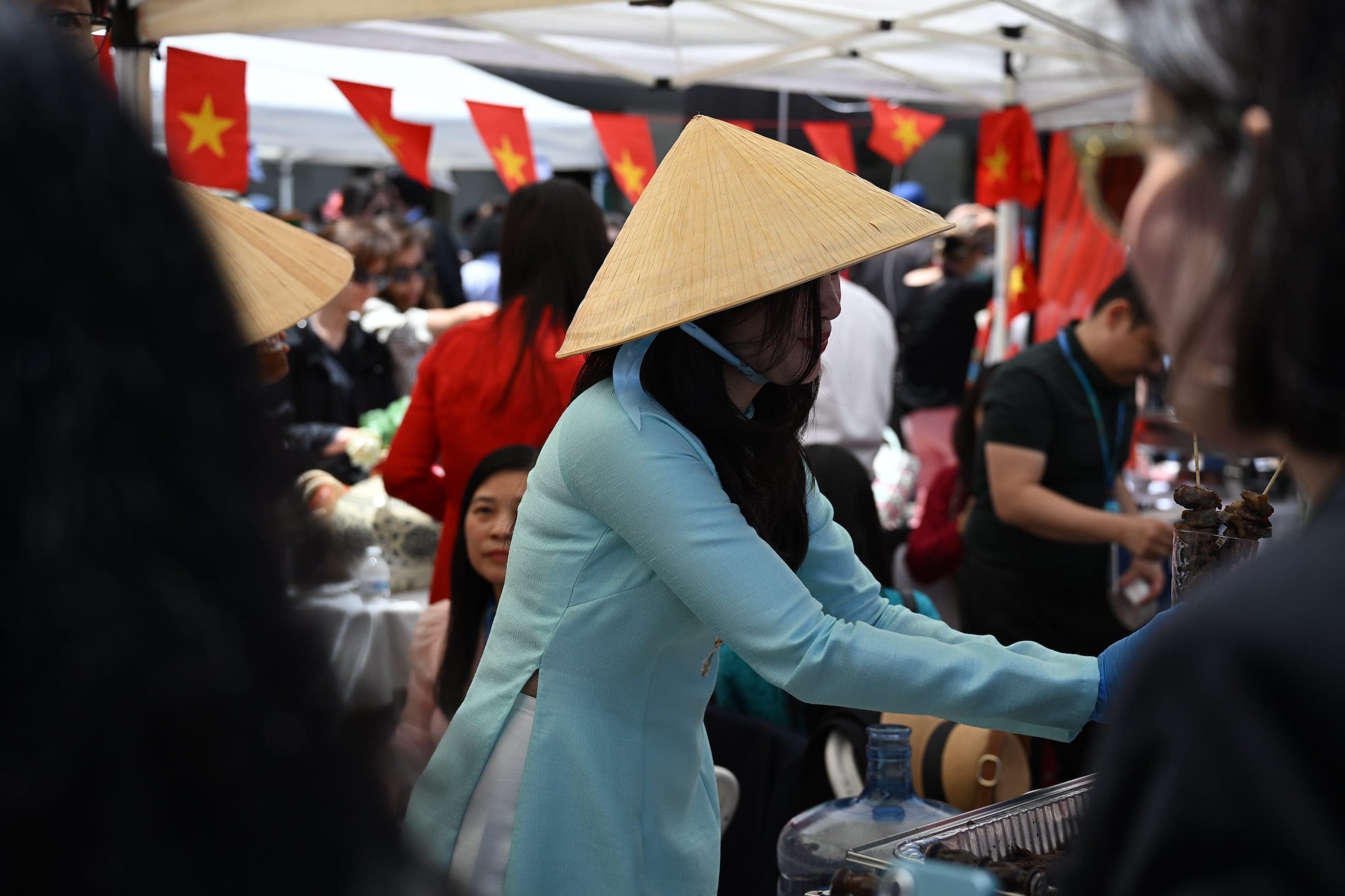 A woman wearing a conical hat serves food beneath Vietnamese flags at an outdoor cultural event.