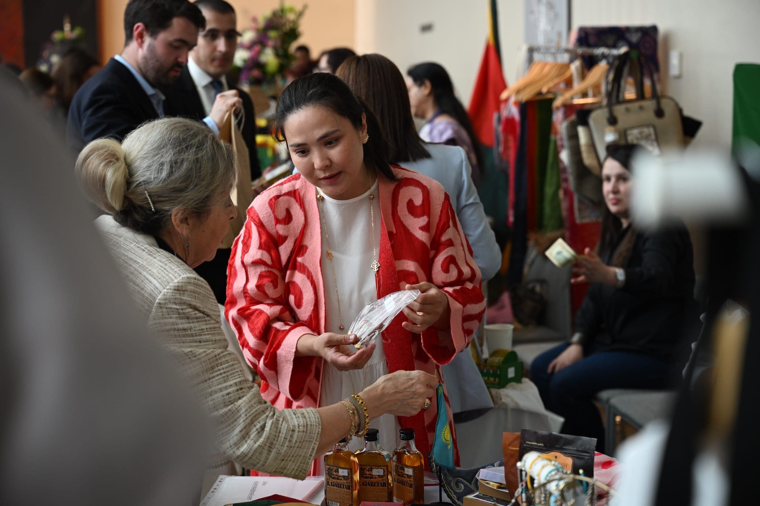 A woman in a red patterned jacket speaks with a visitor at a cultural market stall.