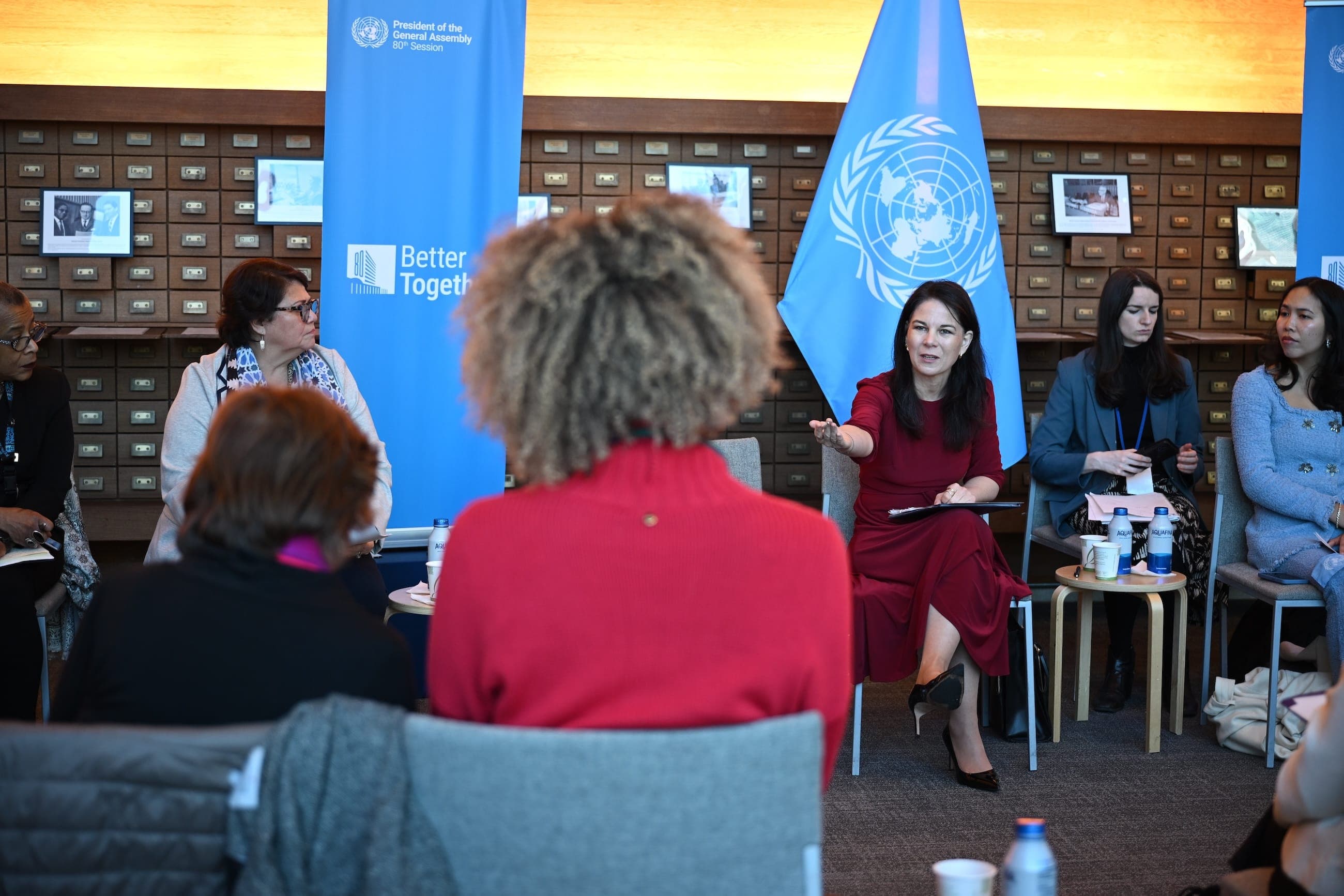 A woman in a red dress leads a round-table discussion before a United Nations flag.