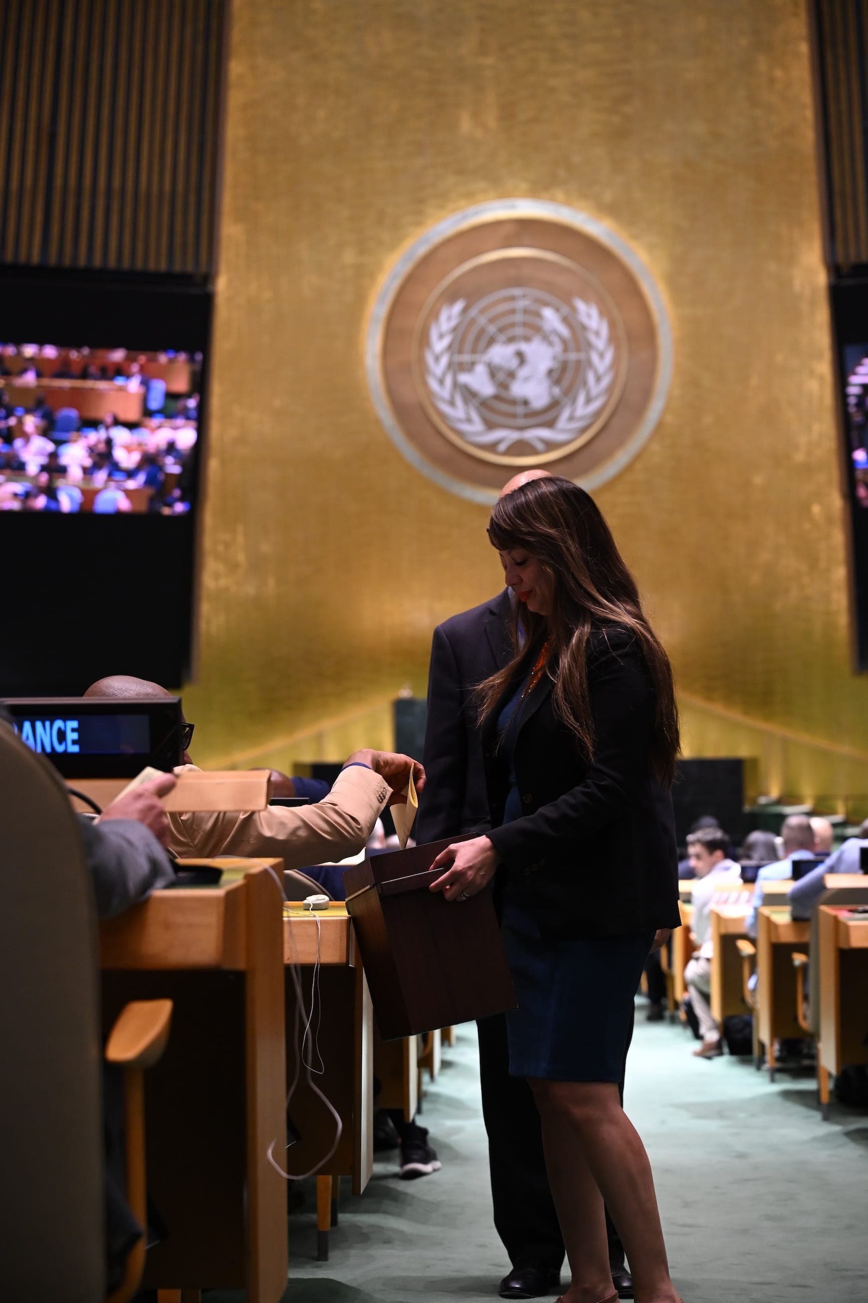 A woman stands at a country desk beneath the United Nations emblem in the General Assembly Hall.