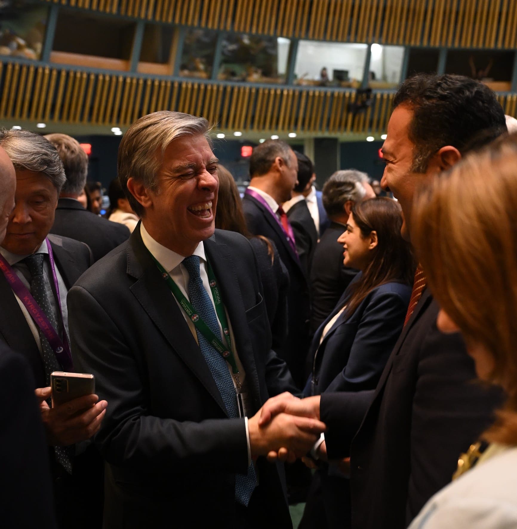 Two men laugh and shake hands among delegates in the General Assembly Hall.