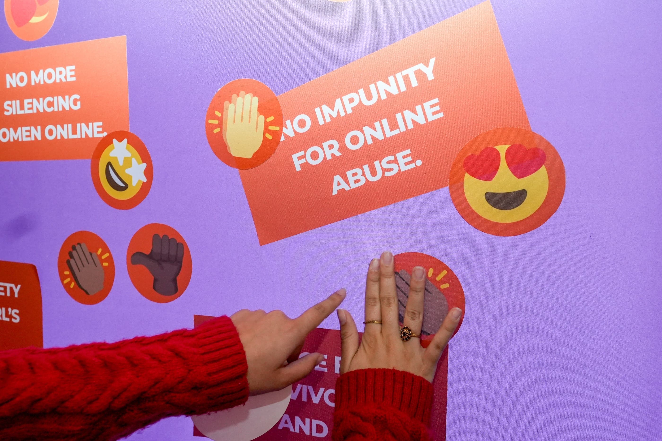 Hands place supportive emoji symbols on the Orange the World installation beside a message calling for no impunity for online abuse.