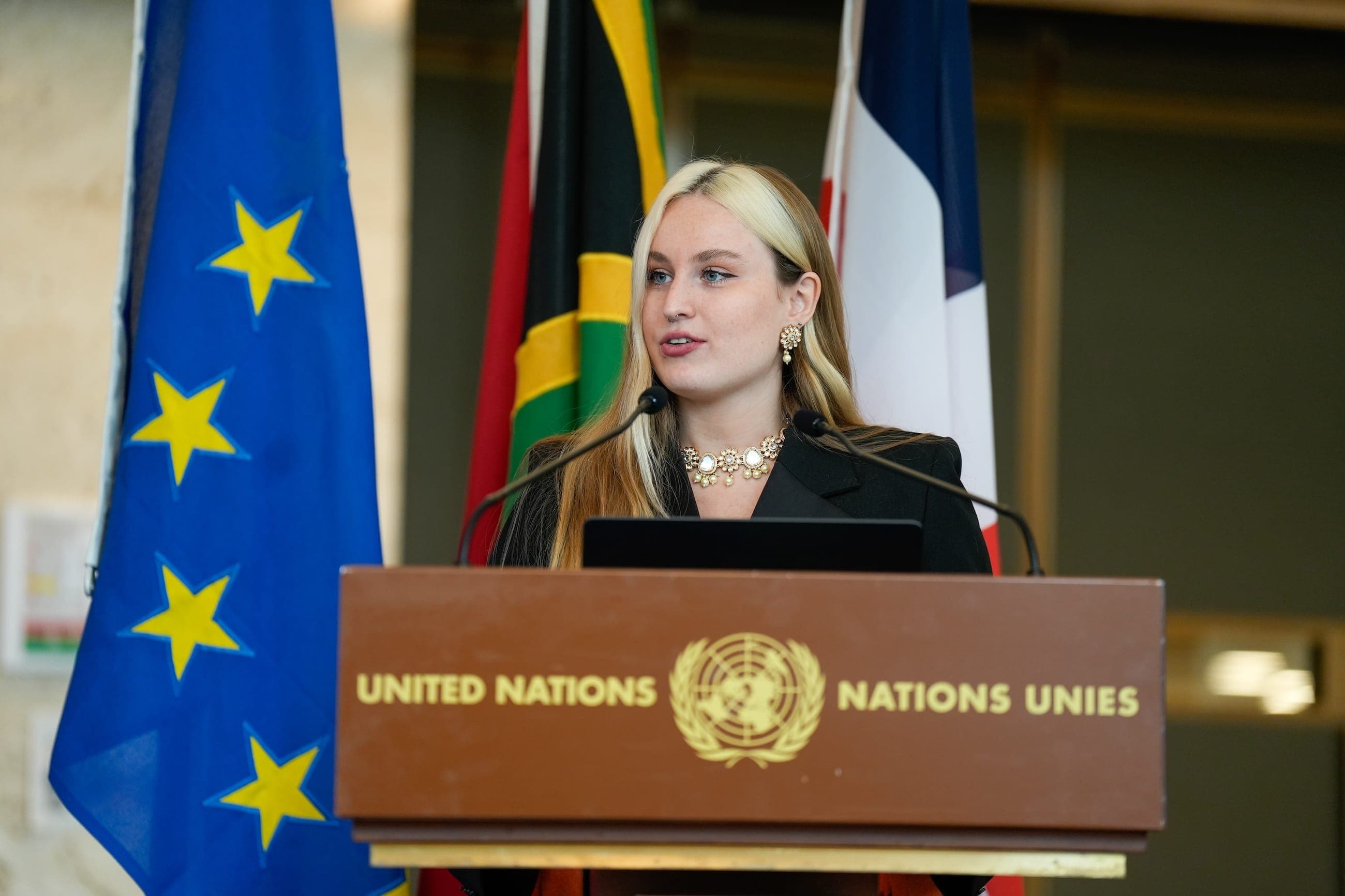 A young woman speaks at a United Nations lectern with European Union and national flags behind her.