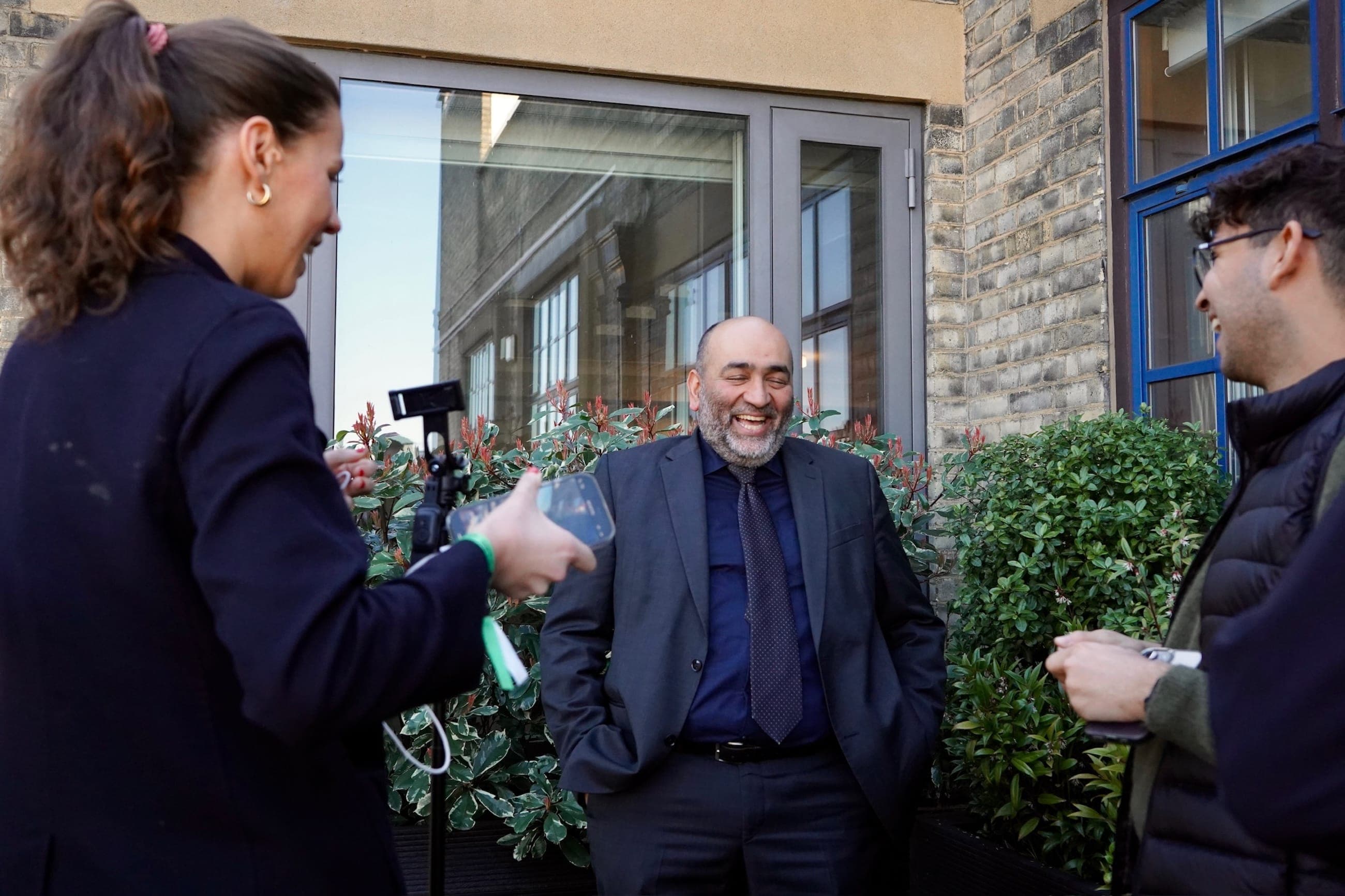 A smiling symposium guest is recorded outdoors by a compact interview team using a smartphone and handheld microphone.