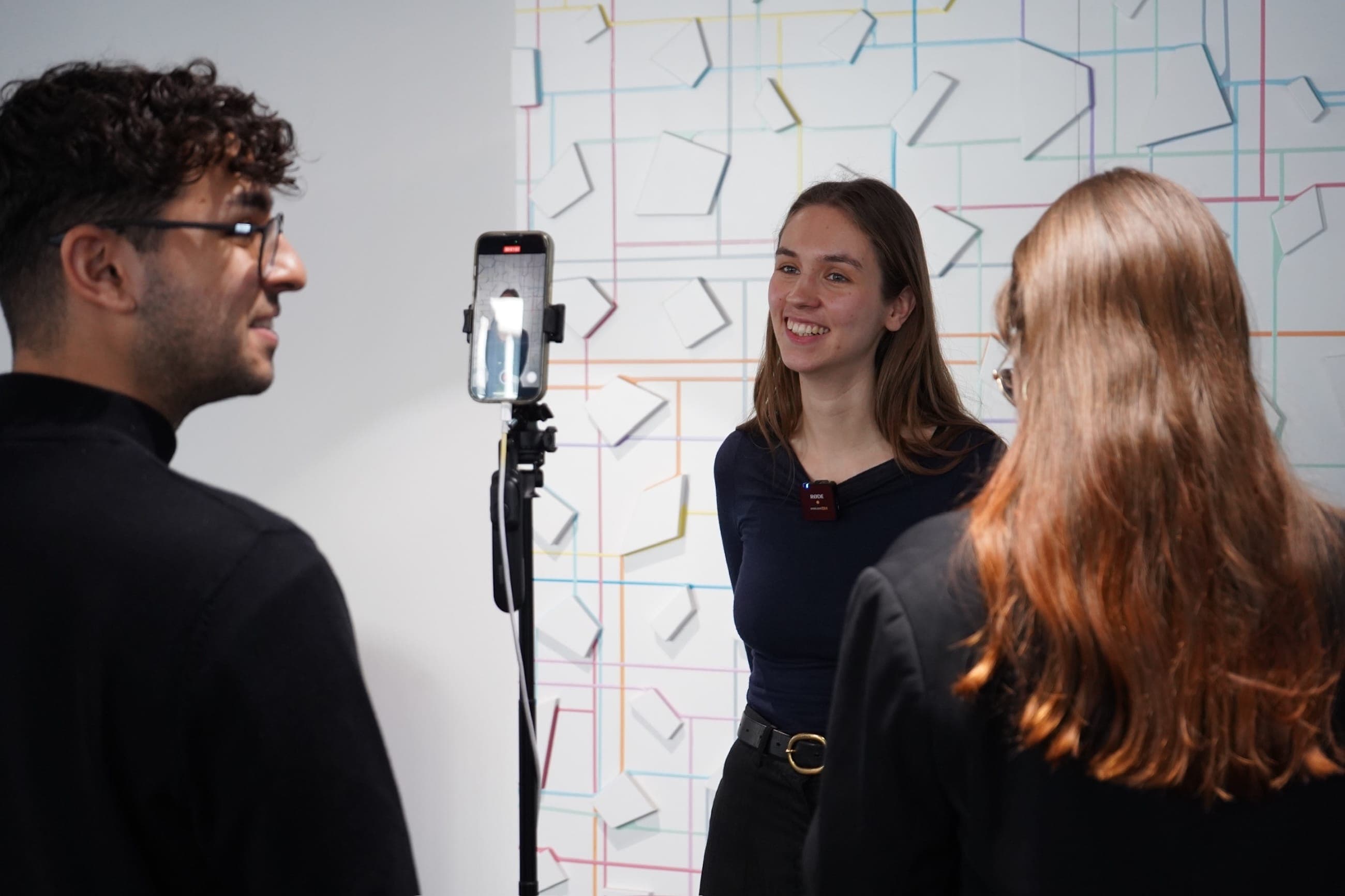 A young symposium speaker wearing a wireless microphone smiles during a smartphone interview as two team members frame the recording.