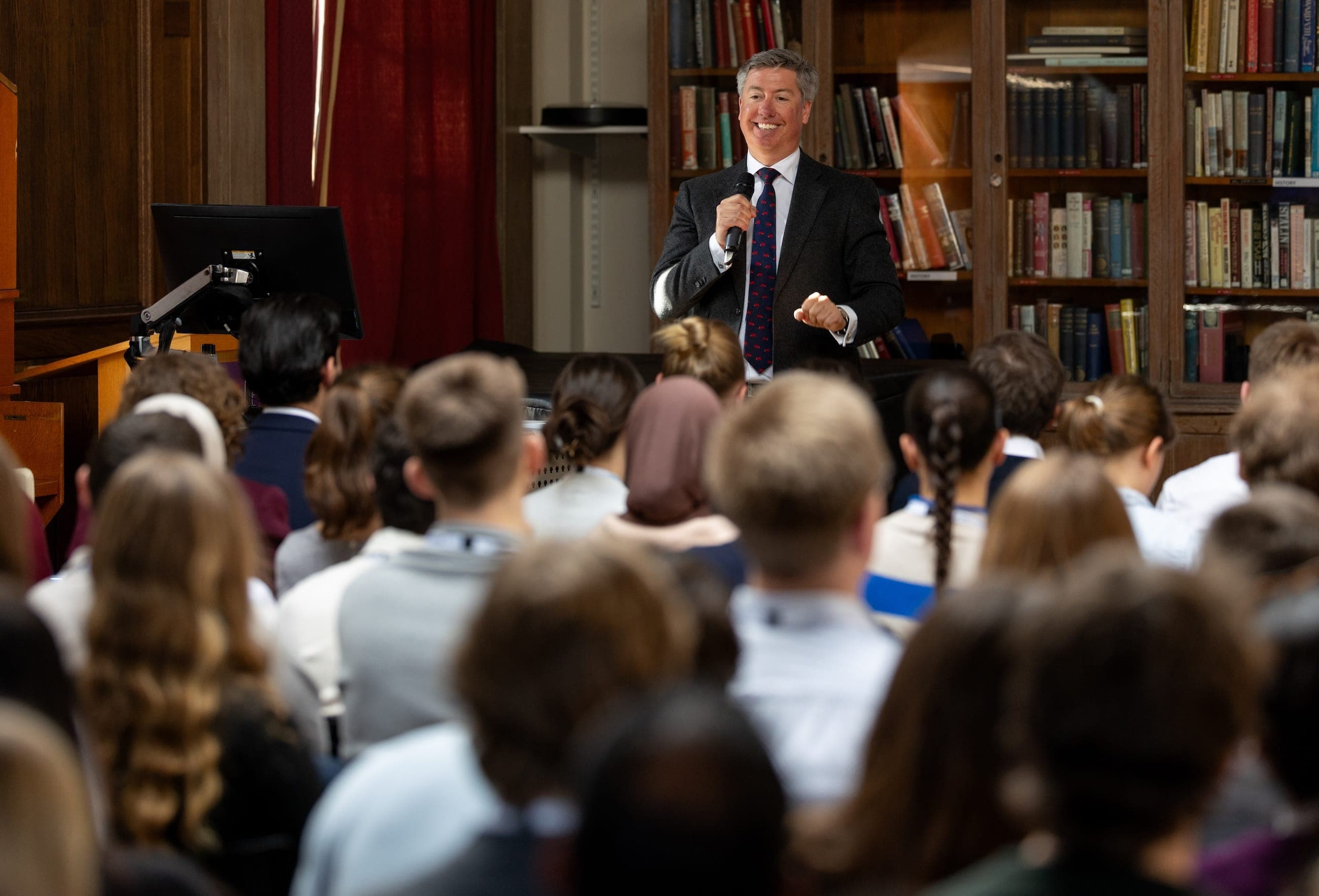 A symposium speaker addresses a seated audience with a handheld microphone in a wood-panelled lecture room.