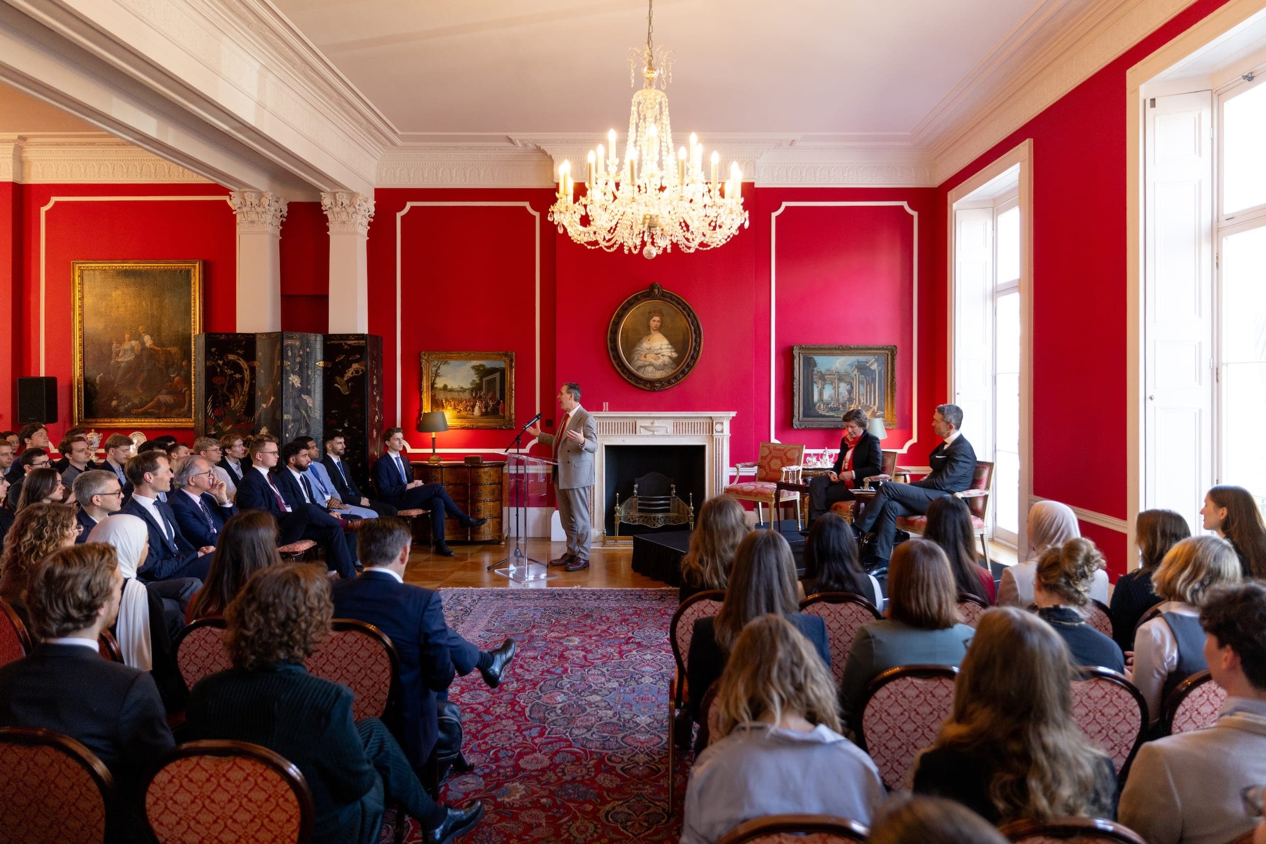 A speaker addresses a large LSE German Symposium audience in a red reception room beneath a chandelier.